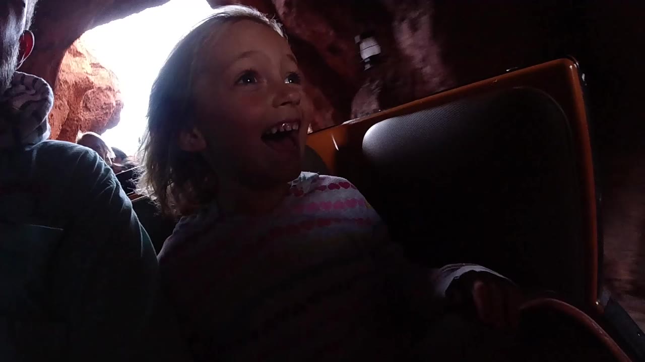 At Disneyland California, a young girl smiles in a dim roller coaster car while bright cave light appears behind nearby riders.