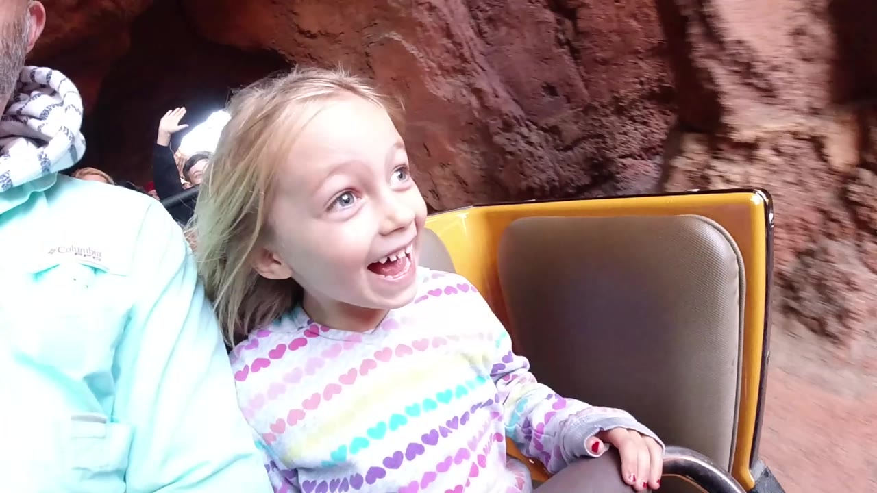 At Disneyland California, a young blonde girl smiles from a yellow ride seat as reddish rock walls fill the background and an adult is cropped at the left edge.