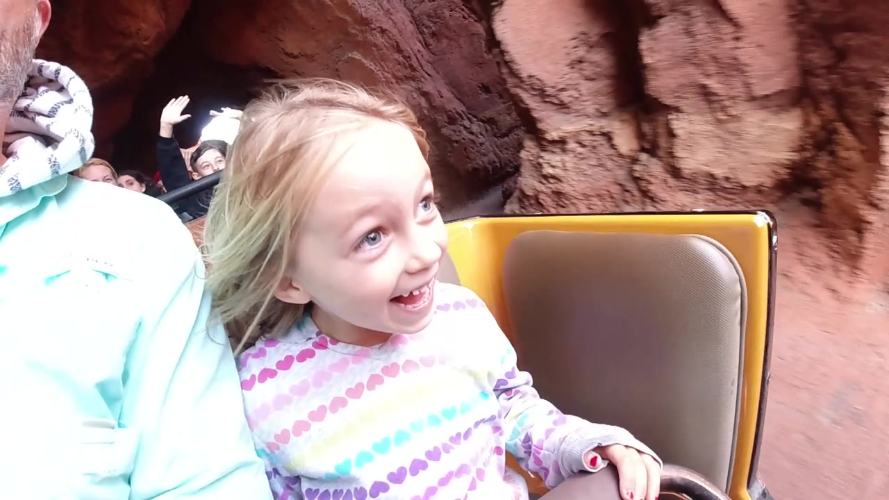 At Disneyland California, the child grins toward the right side of frame as the ride car passes close to reddish rock scenery and raised hands appear behind her.