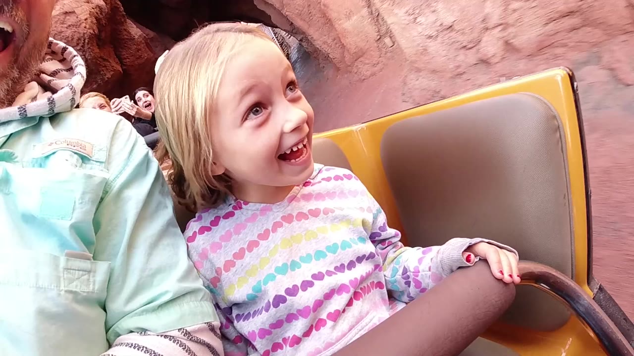 At Disneyland California, a blond young girl smiles wide in a yellow roller coaster car while seated beside an adult in a mint shirt, with red rockwork behind them.