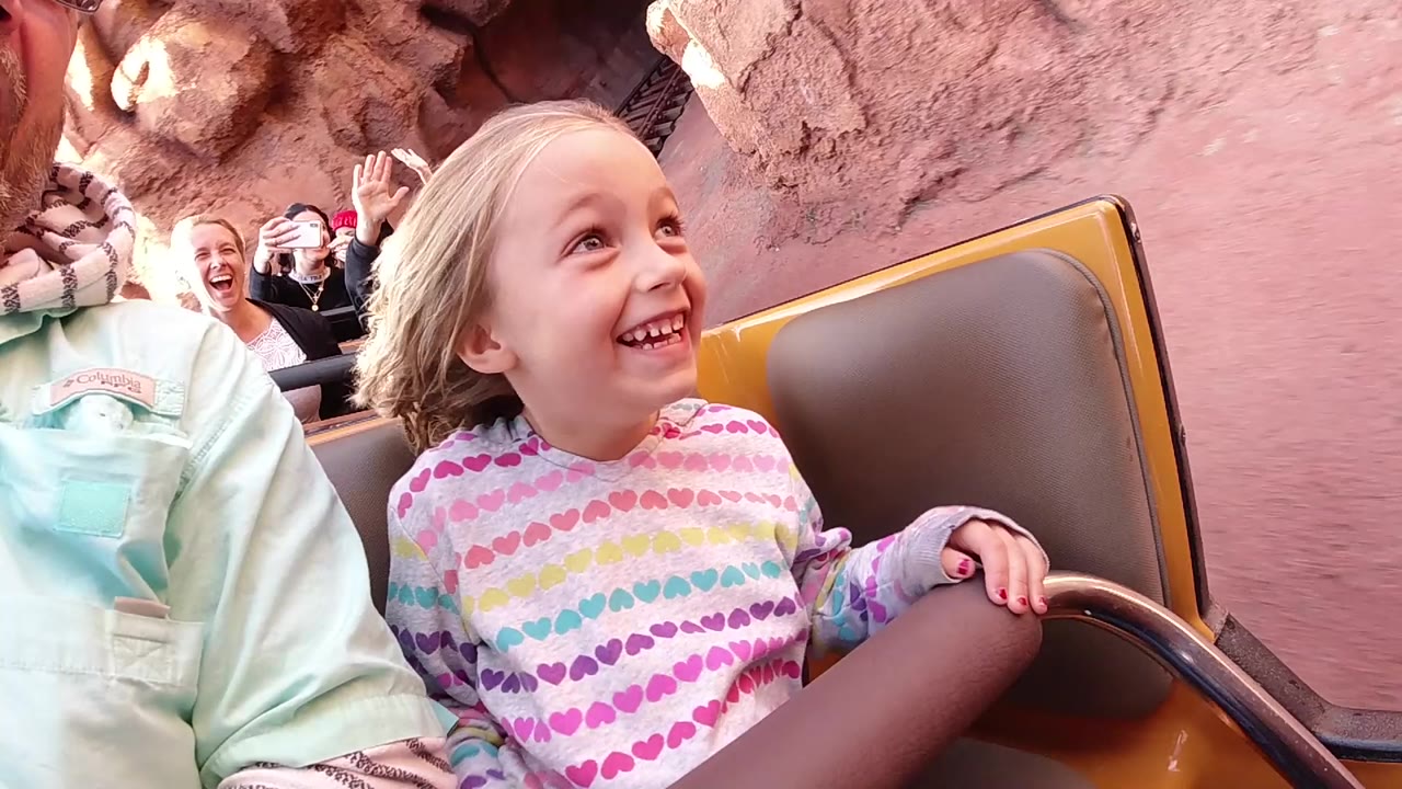 At Disneyland California, the girl grins toward the side of the roller coaster car as adults behind her smile and raise hands near the rock-lined track.