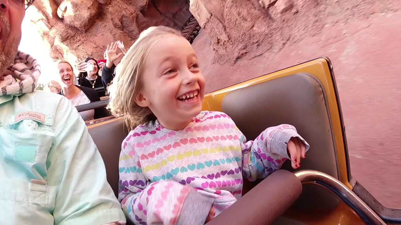 At Disneyland California, the girl laughs broadly in the roller coaster car, with one hand on the seat edge and adults visible at the sides and behind her.