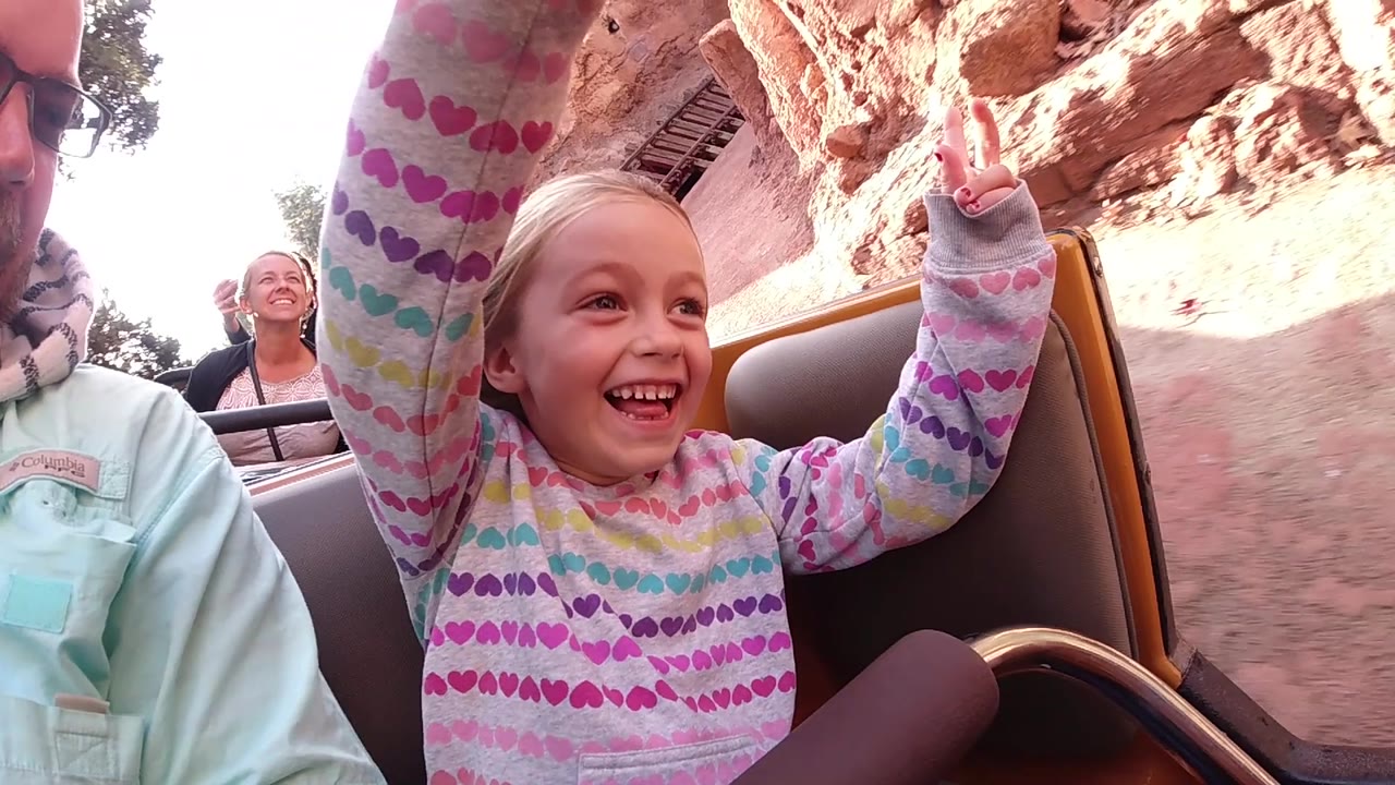 At Disneyland California, the girl grins and looks to the side with her arms up, while a smiling adult rider appears in the row behind and rockwork passes along the right side.