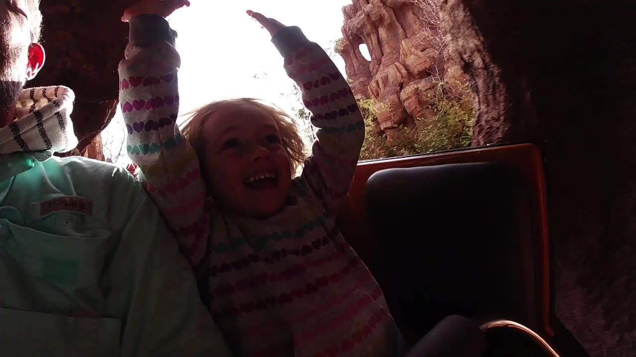 At Disneyland California, a blond child raises both arms beside an adult man in a ride car, with dark rockwork framing a bright opening behind them.
