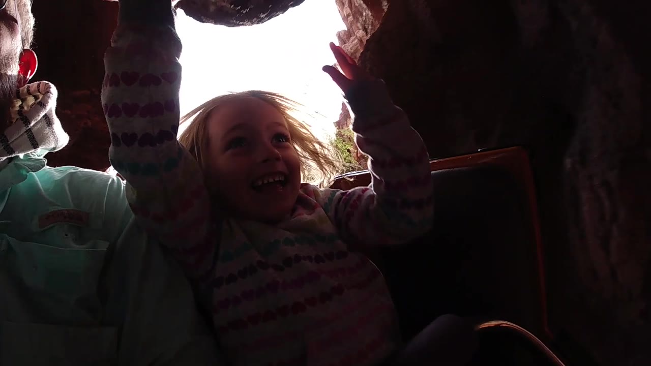 At Disneyland California, the child laughs and looks upward with one arm raised, while the adult beside her is partly silhouetted by daylight behind the rock opening.