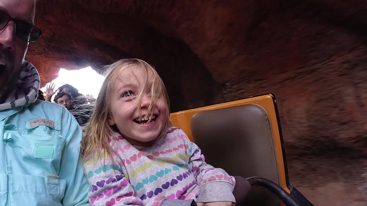 At Disneyland California, a young girl laughs beside an adult man in sunglasses as their rollercoaster car passes through a reddish rock tunnel.
