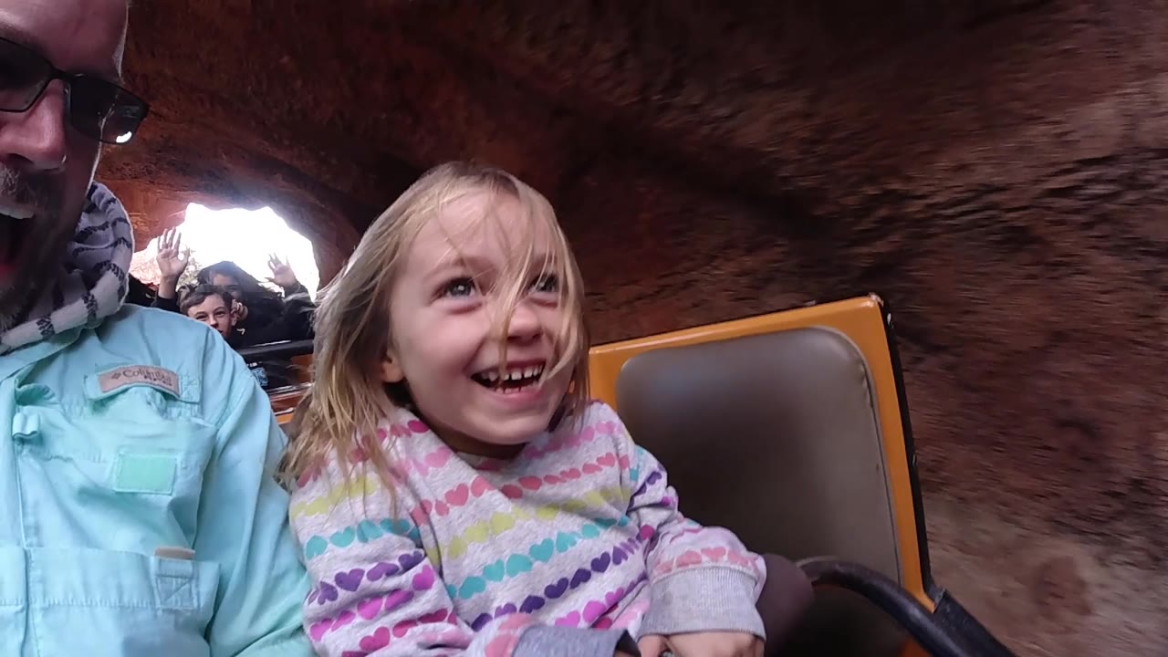The girl grins with hair across her face while the adult man sits partly cropped beside her and the textured reddish ride wall fills the background at Disneyland California.