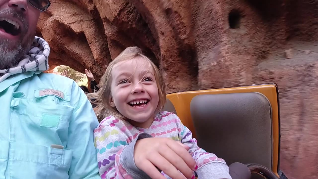 At Disneyland California, a young blonde girl smiles in a close ride-vehicle view while a cropped adult in a light shirt sits beside her and reddish rockwork fills the background.