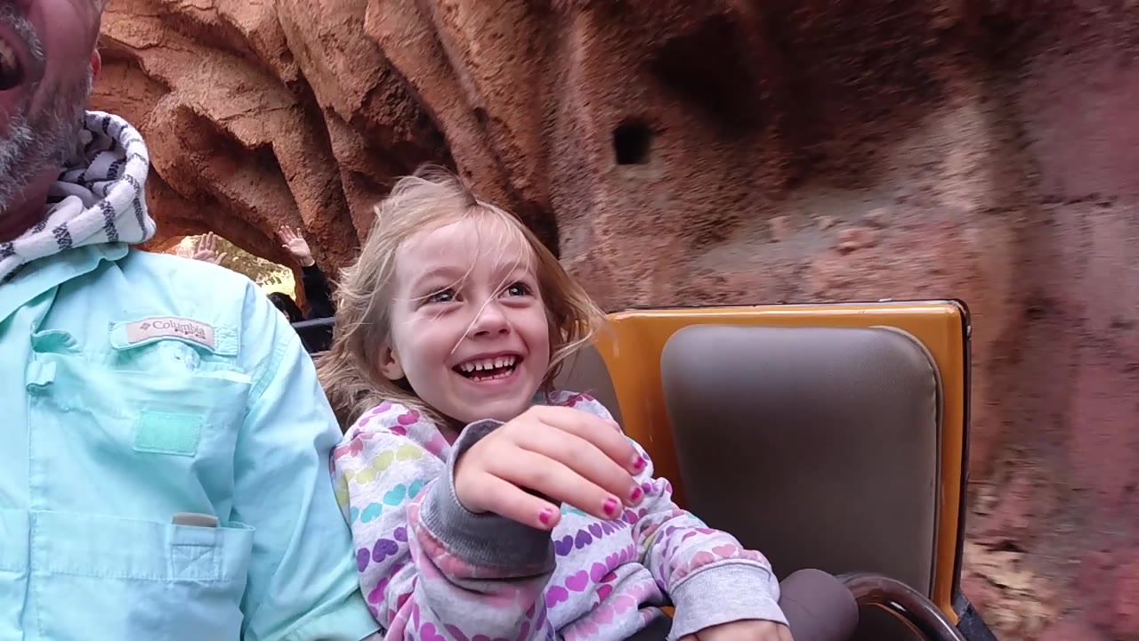 At Disneyland California, the child smiles while extending one hand forward, with a brown seat back beside her and reddish artificial rock texture behind the ride vehicle.