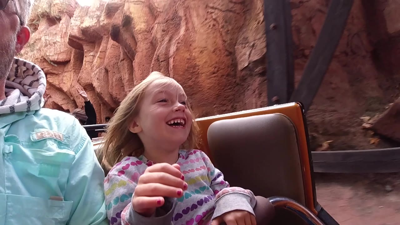 At Disneyland California, the child laughs with one hand raised near the lap area while red rock walls and a dark rail edge run behind the coaster car.