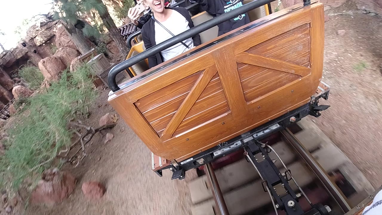 At Disneyland California, the coaster car remains close to camera as riders sit behind the rail, with reddish rocks, shrubs, and track hardware visible below.