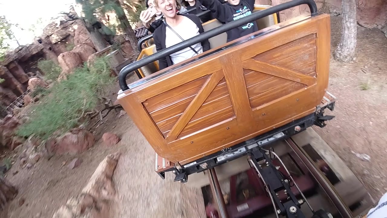 At Disneyland California, the coaster car is angled across the frame with riders partly cropped above the rail and rocky terrain and ride track visible below.