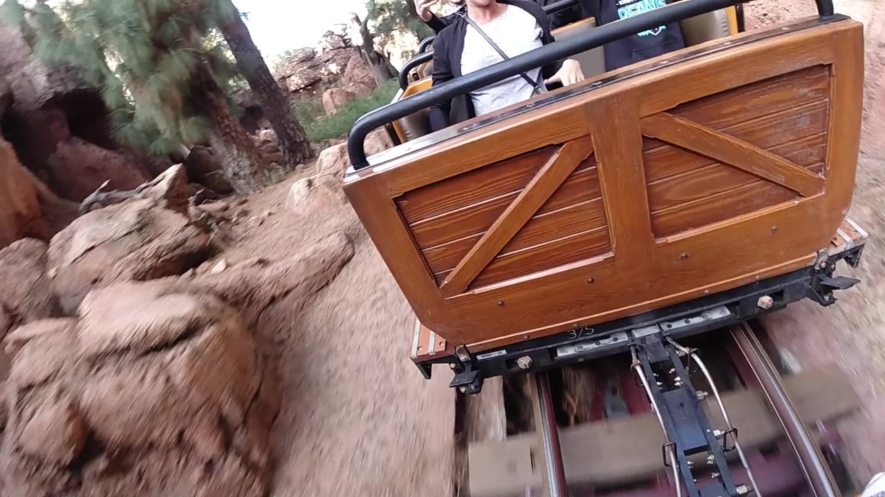 A wood-paneled roller coaster cart rides along metal rails at Disneyland California, with reddish rock walls, trees, and partial riders visible above the front rail.