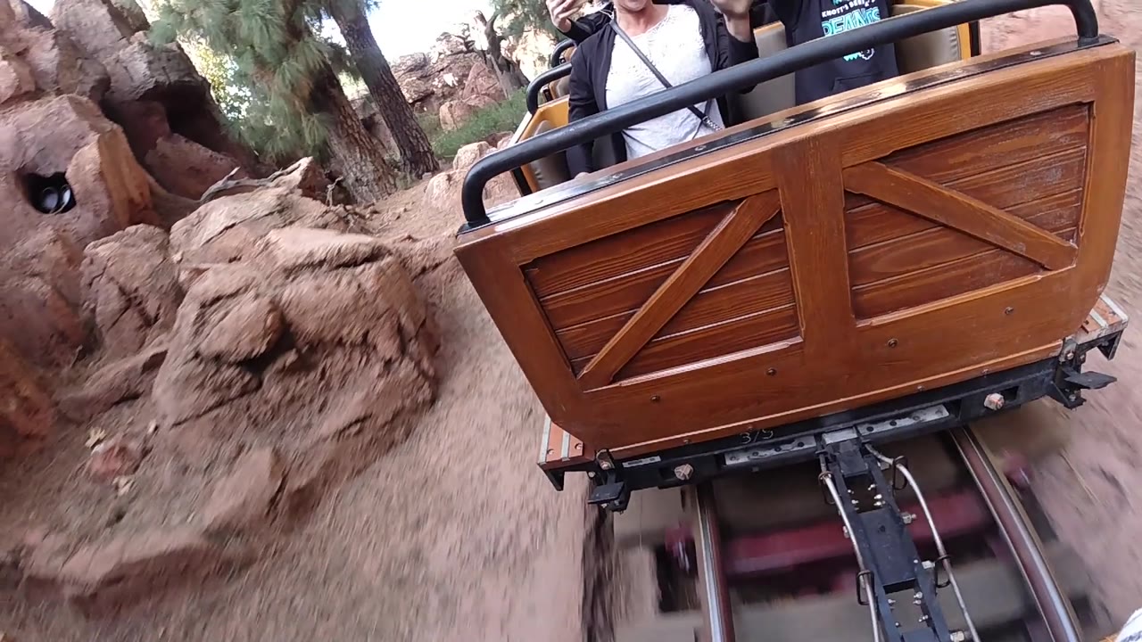 A close view of the cart at Disneyland California shows wood paneling, black railings, exposed track hardware, reddish rocks, trees, and partial riders in the upper frame.