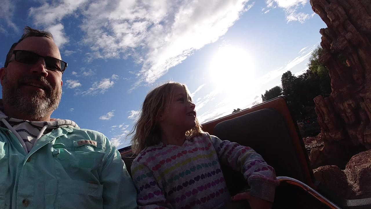 At Disneyland California, a dad in sunglasses and a young daughter sit in a ride vehicle with rockwork at the edge of frame and bright sun behind them.