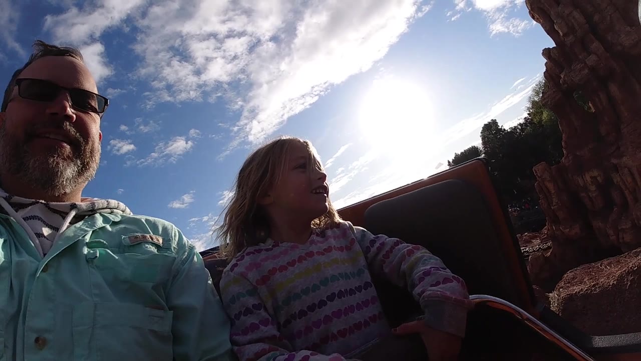At Disneyland California, the seated pair are backlit against blue sky and white clouds, with the daughter's patterned sweater and the ride seat visible in the foreground.