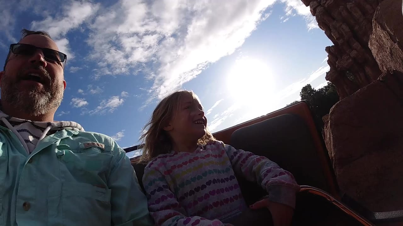 At Disneyland California, a father in sunglasses and a young girl sit close to the camera in a roller coaster car with rockwork, blue sky, and strong sun behind them.