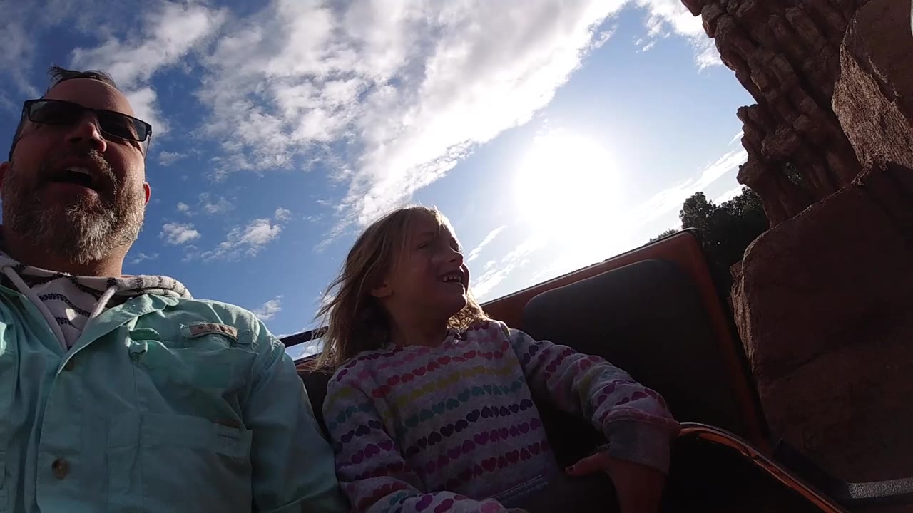 At Disneyland California, the father remains at the left edge while the girl looks toward the rock wall, with the sun high behind the coaster car.