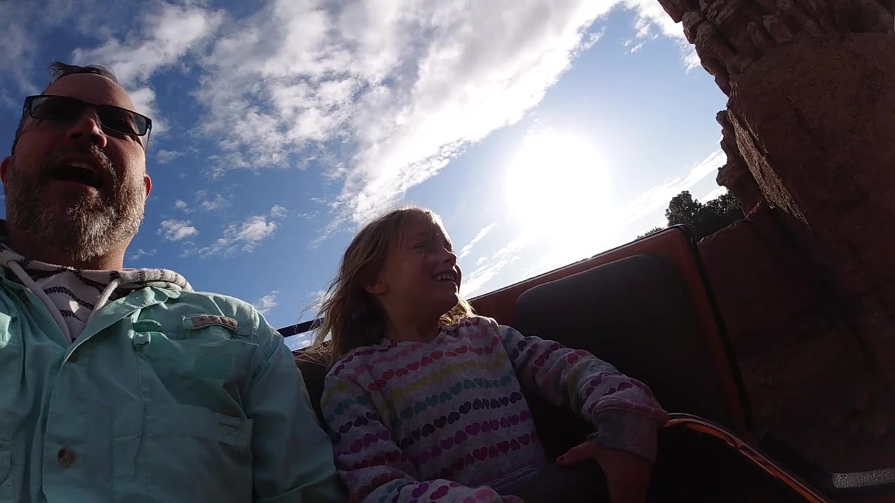 At Disneyland California, the girl smiles toward the side of the ride while the father sits beside her, backlit by the sun and framed by red rock structures.