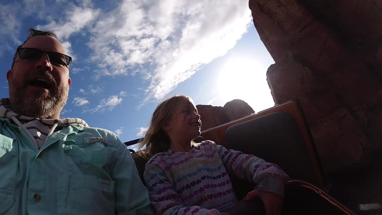 At Disneyland California, a bearded man in sunglasses and a young girl sit in a ride vehicle as bright sun and reddish rockwork fill the background.