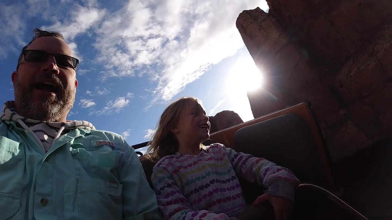 At Disneyland California, the riders are seen from a low close angle as the sun flares near the edge of the reddish rockwork behind the girl.