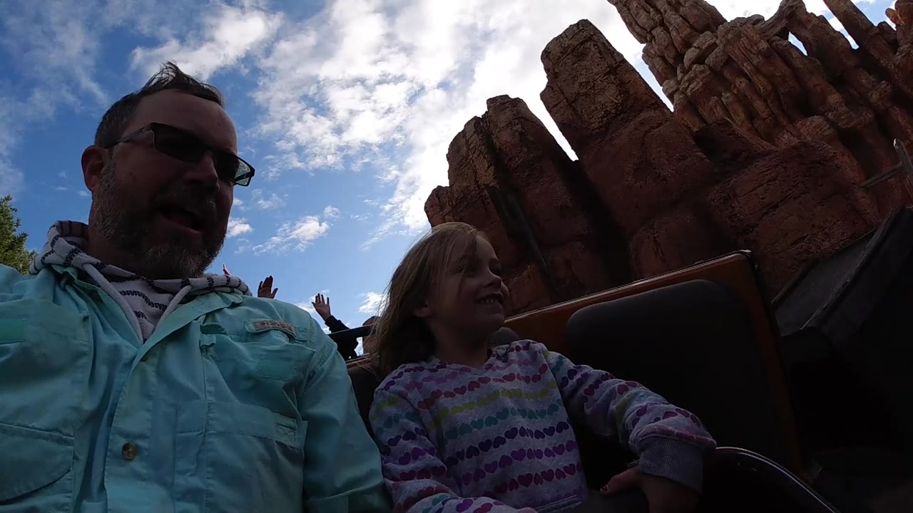 At Disneyland California, a father in sunglasses and a young girl sit in the foreground of a roller coaster car with red canyon-style rock formations and blue sky behind them.