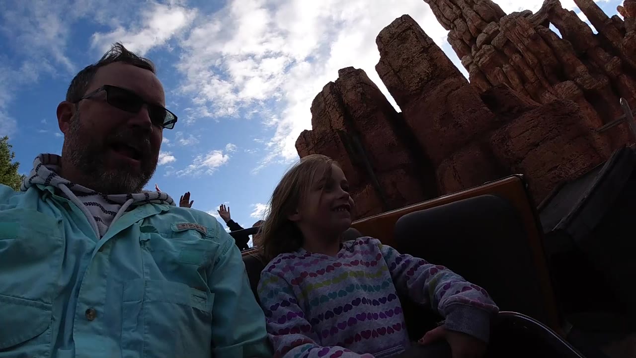 At Disneyland California, the father and daughter remain seated close to camera while jagged red rockwork fills the upper right of the frame under scattered clouds.