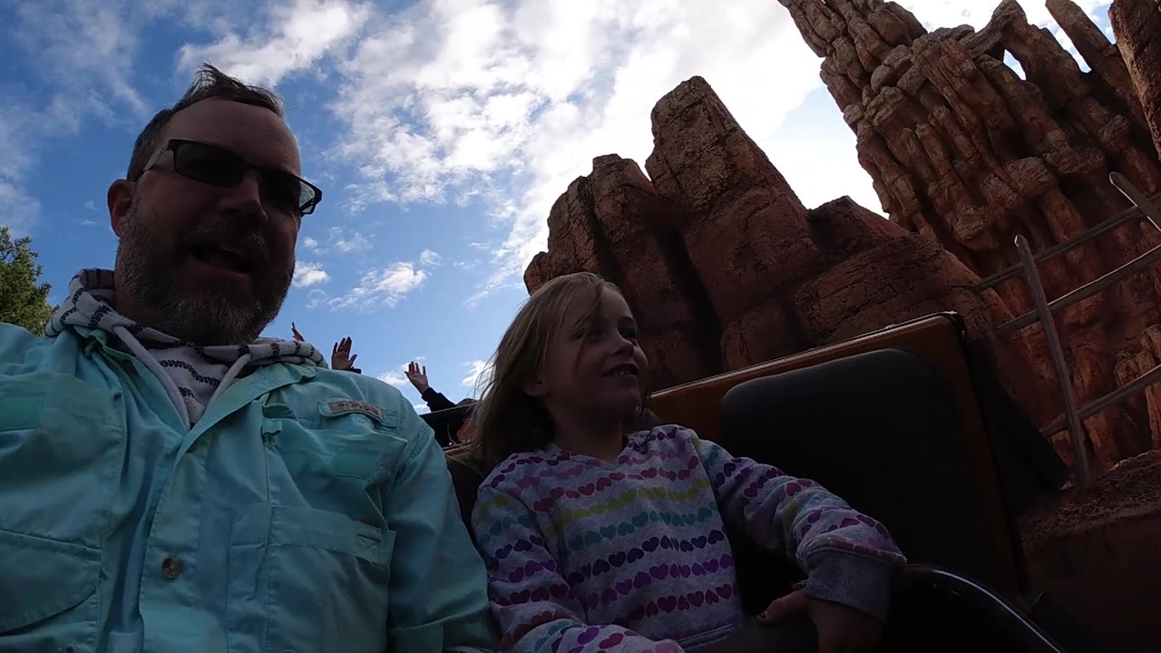 At Disneyland California, the father looks toward the ride path while the young girl sits beside him, with tall red rock forms and a bright cloud bank behind the coaster car.