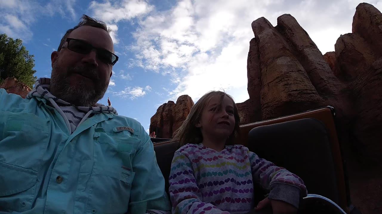 At Disneyland California, an adult in sunglasses and a child sit in a roller coaster car under a bright cloudy sky, with reddish rock formations behind them.