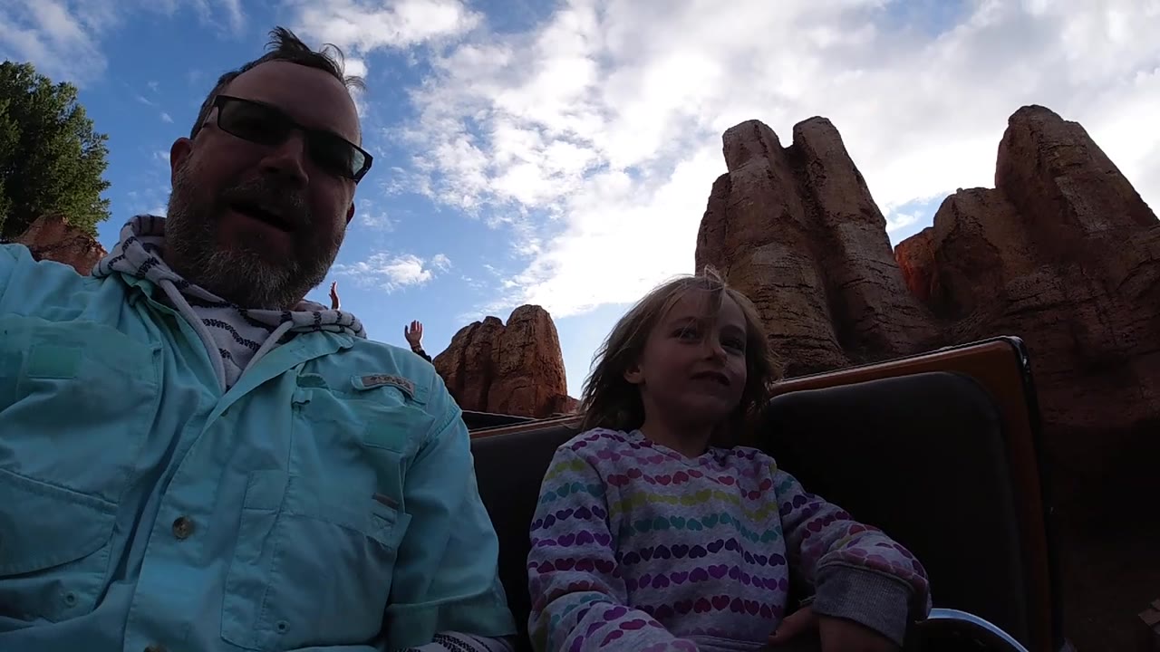 At Disneyland California, the adult and child sit in the coaster car with a raised hand from another rider visible between the reddish rock formations.
