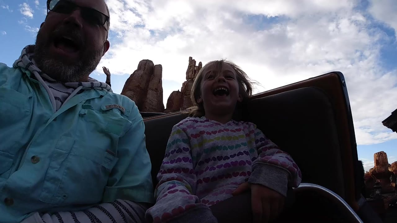 At Disneyland California, a father in sunglasses and a child in a patterned sweater laugh in a dark backlit roller coaster seat with rockwork behind them.