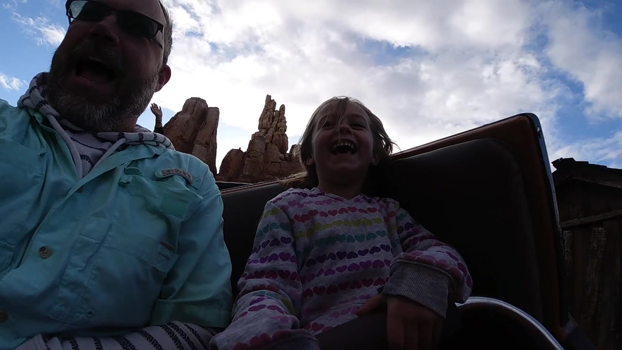 A backlit Disneyland California coaster moment shows the father cropped along the left edge and the child smiling beside a dark seat side under bright clouds.