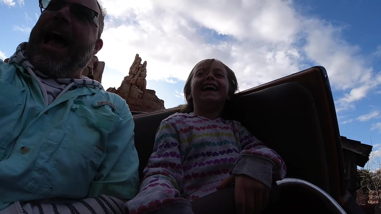 Child laughing beside an adult on a roller coaster with clouds and rockwork behind.