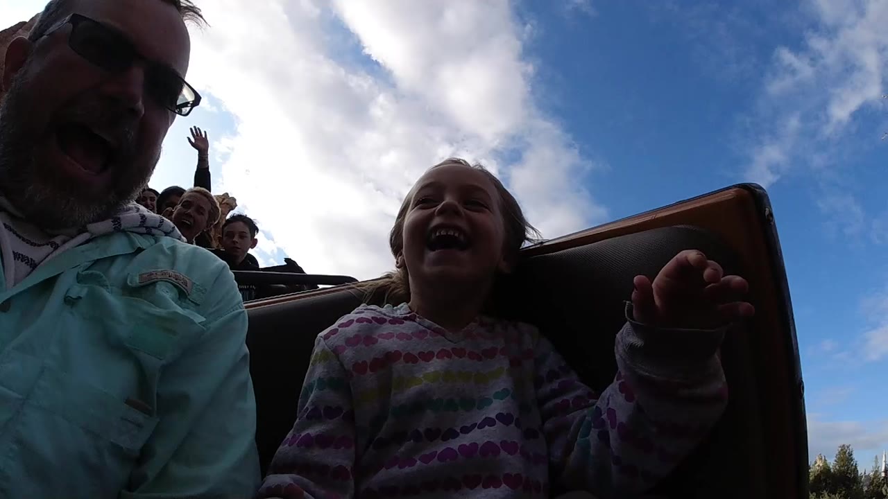 Young girl laughs beside her father with riders visible behind them.