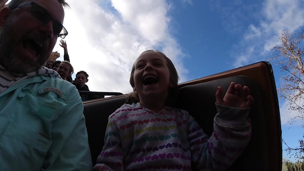 At Disneyland California, the young girl keeps laughing with one hand lifted near the seat edge, while the adult man and background riders remain partly in shadow.