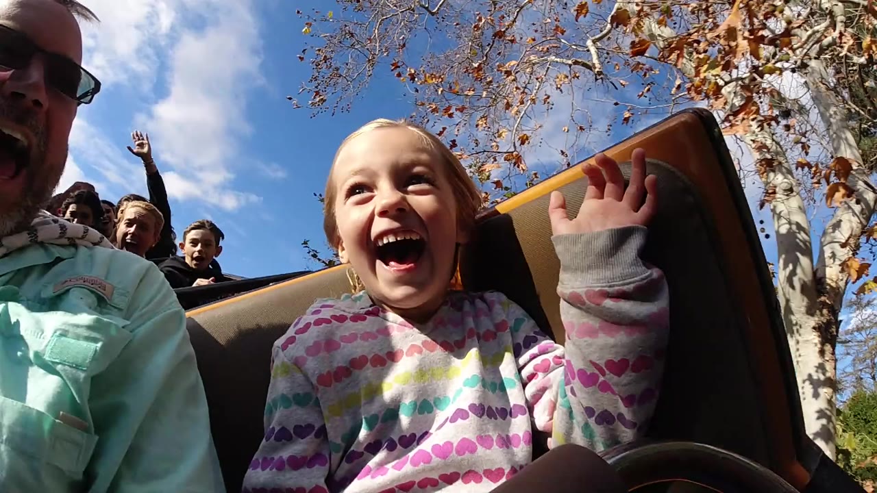 At Disneyland California, a young girl laughs in the foreground beside an adult while other riders sit behind her under blue sky and tree branches.