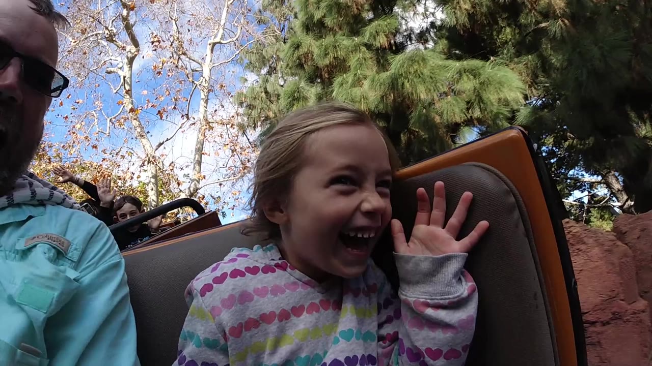 At Disneyland California, a young girl laughs with her hand raised in a ride seat while an adult in sunglasses is partly visible at the left edge.