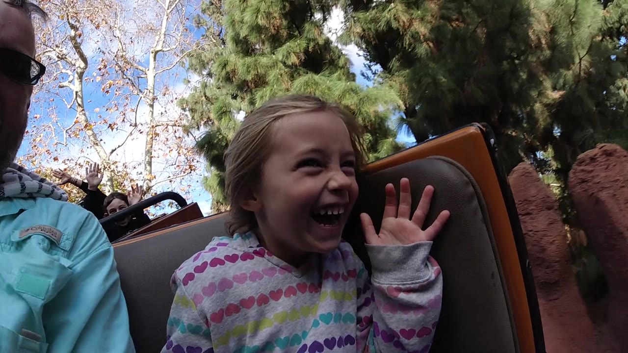 At Disneyland California, the laughing girl sits between a partially visible adult and a dark ride seatback, with red rockwork appearing closer on the right.
