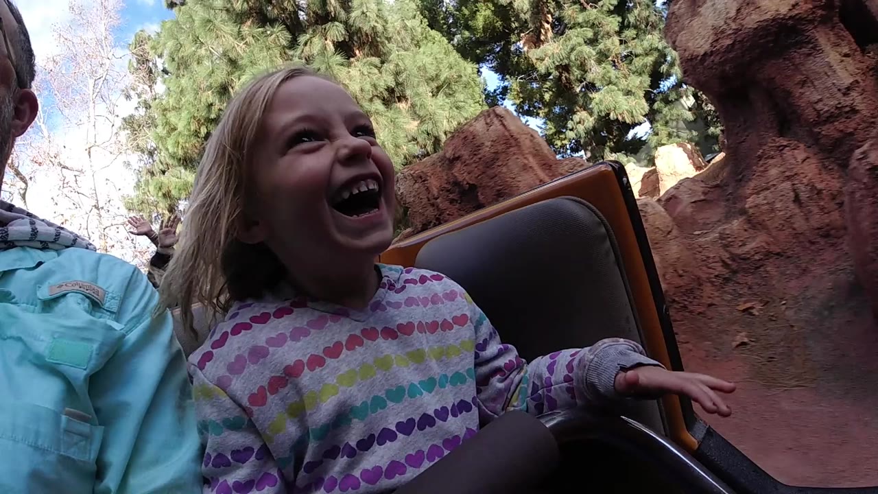 At Disneyland California, a laughing young girl in a white multicolor striped shirt sits beside a cropped adult with red rock scenery and trees behind her.