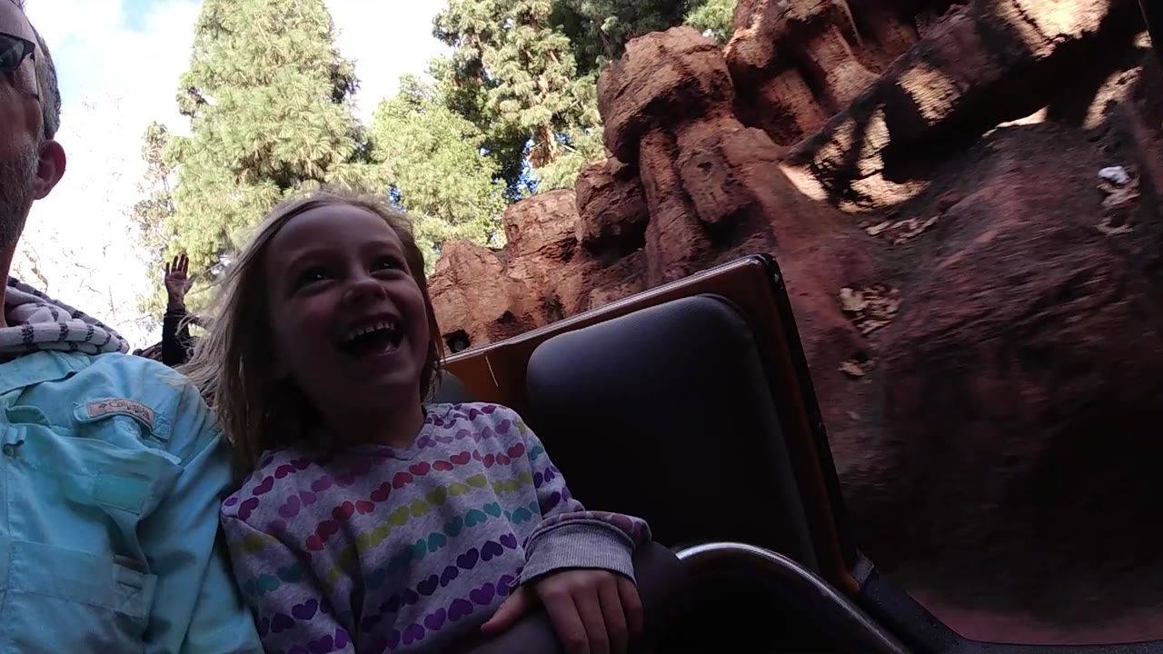 At Disneyland California, the girl remains centered and smiling while the adult's shoulder and face edge frame the left side and red rock-like scenery fills the background.