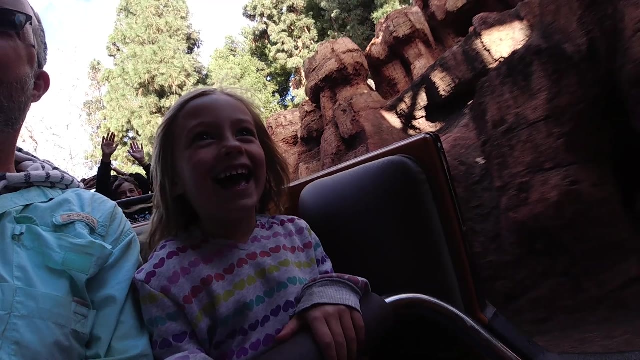 At Disneyland California, the girl laughs in the foreground while a rider in the background raises both hands between the coaster seats and rock-like walls.