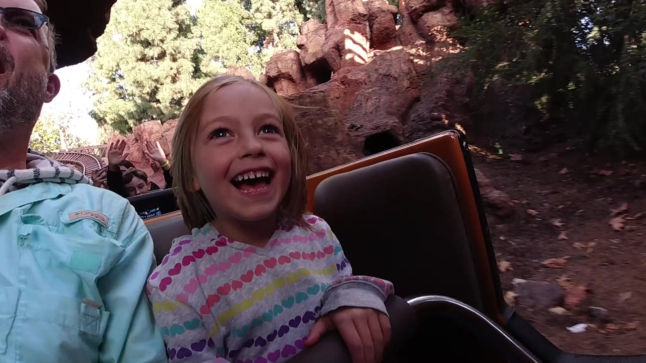 At Disneyland California, the child laughs while seated next to a cropped adult, with another rider faintly visible behind and red rock scenery in the background.