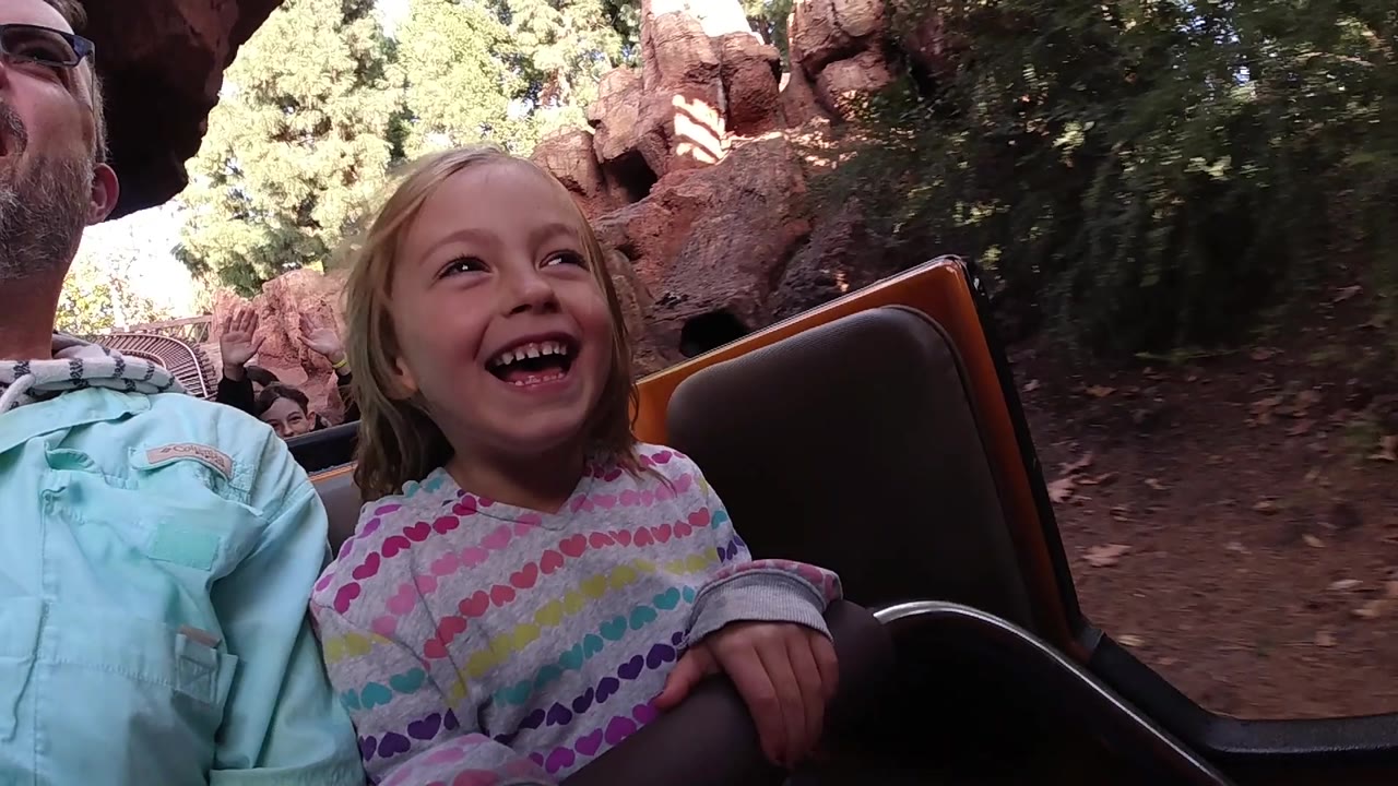 At Disneyland California, the girl looks upward with a broad smile as the ride car passes trees and red rock-like structures in mixed sunlight.