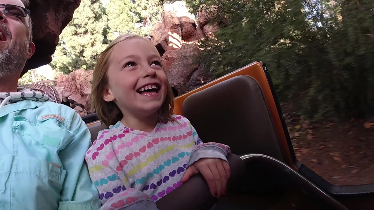 Child grinning in a brown ride seat while an adult is cropped at left.