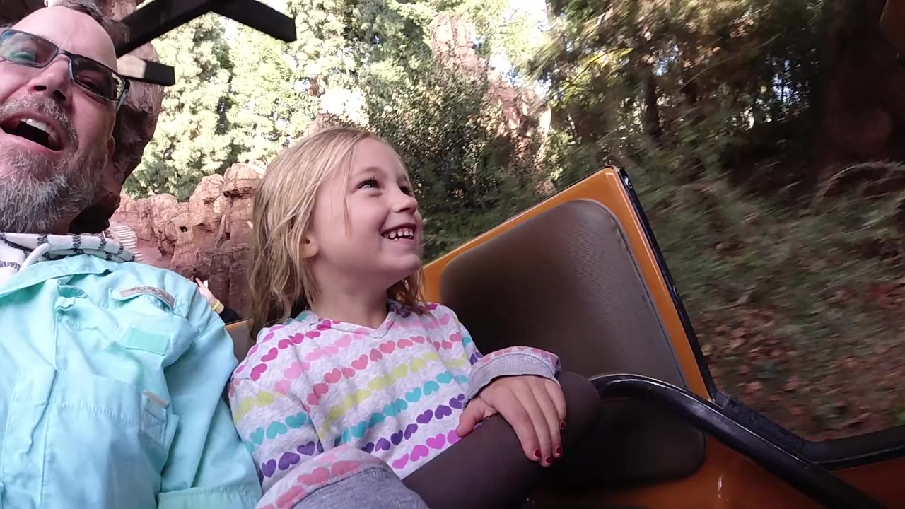 At Disneyland California, a father in glasses and a blonde young daughter smile in the front of a roller coaster car with trees and red rock scenery behind them.
