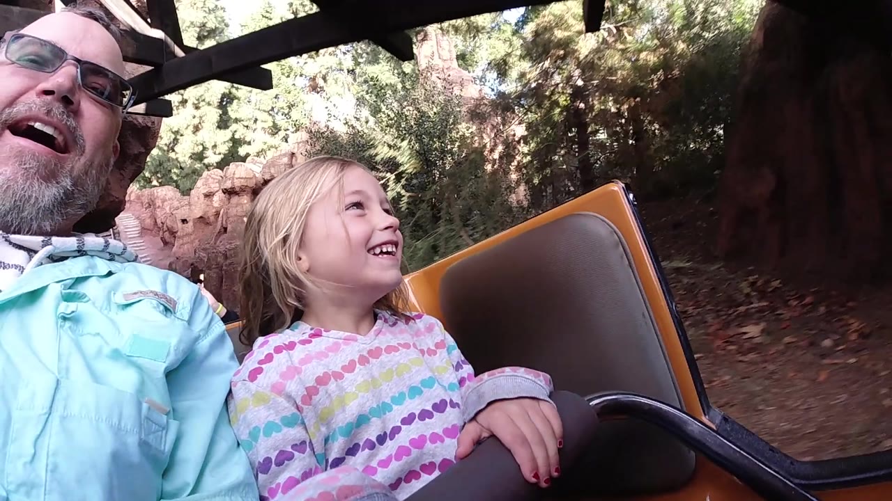 The father and daughter look up and laugh from the coaster car as darker ride structure and shaded rockwork appear around the Disneyland California track area.