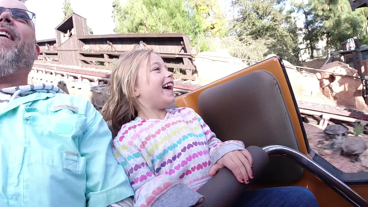 At Disneyland California, a blonde girl laughs beside a partly cropped older man in sunglasses while seated in a brown amusement ride car with trees and wooden structures behind them.