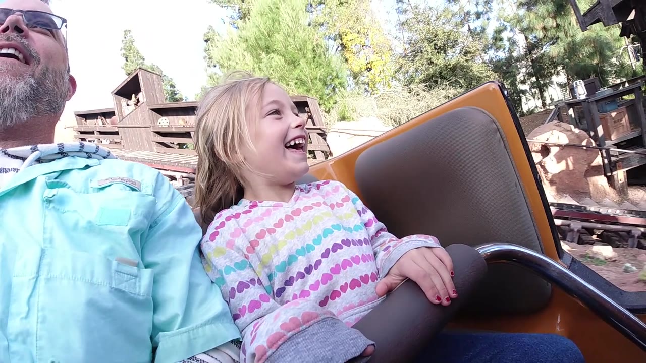 At Disneyland California, the child smiles broadly while gripping the ride car edge, with the grandfather partly visible at left and a rustic wood building and trees behind the track area.