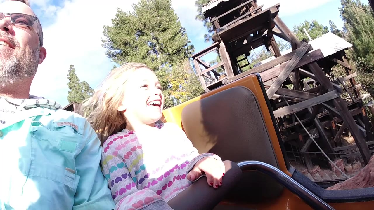 At Disneyland California, an adult man and young girl sit close to the camera in a coaster car as wooden ride structure, trees, and blue sky fill the background.
