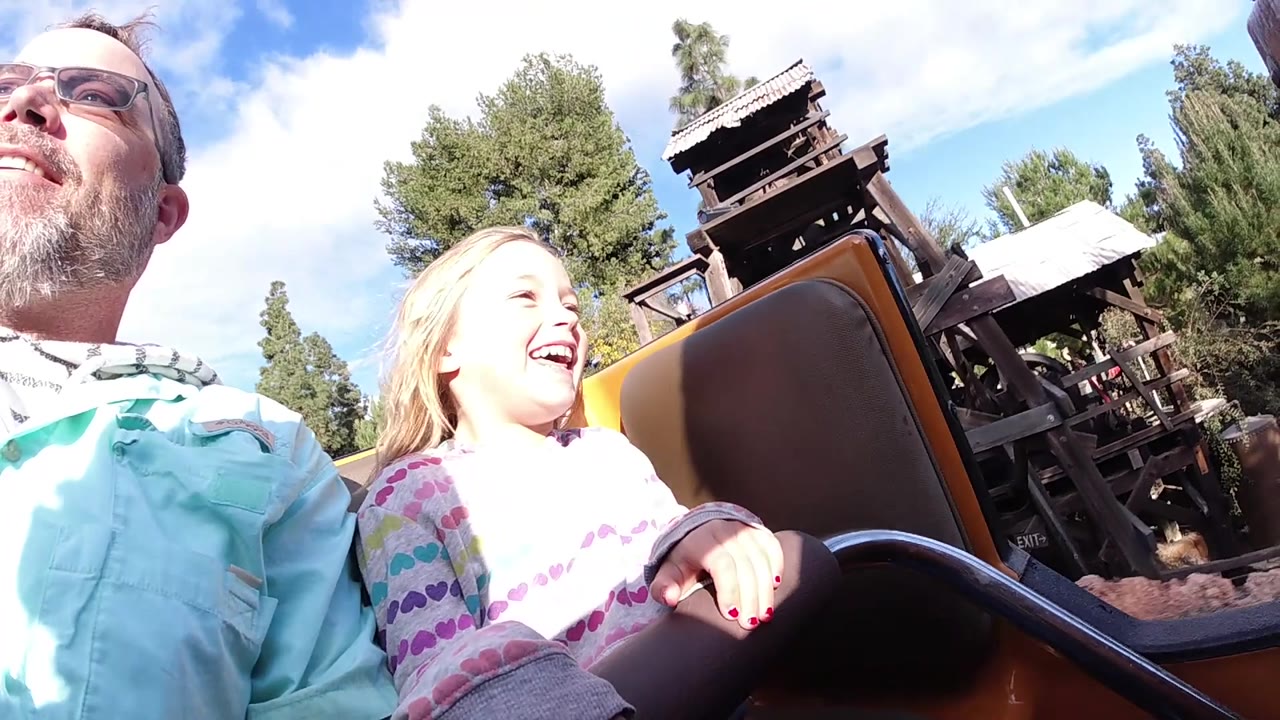 At Disneyland California, the girl smiles broadly in the foreground while the brown coaster seat and timber ride elements angle through the background.